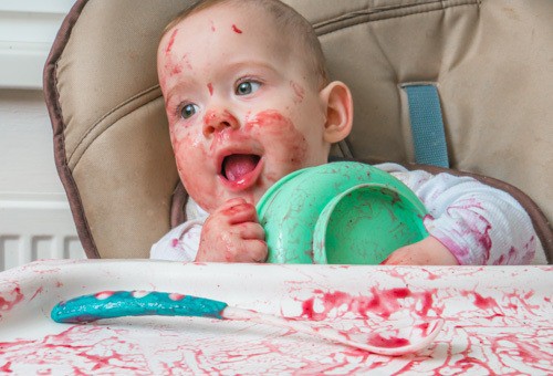 Messy little baby is eating dinner from bowl.